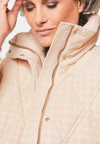 Beige gesteppte Jacke mit hohem Kragen, gerippter Textur und dezentem Muster. Verfügt über einen Reißverschluss und Druckknopfverschlüsse.