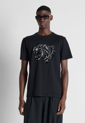 Slanke man met zwarte zonnebril en een zwart t-shirt met een afbeelding van een brullende luipaard aan de voorkant, tegen een grijze achtergrond.