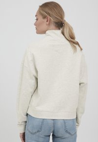 Lys grå, tekstureret sweatshirt med høj krave og dropped skuldre, parret med lys blå denim jeans, set fra ryggen.
