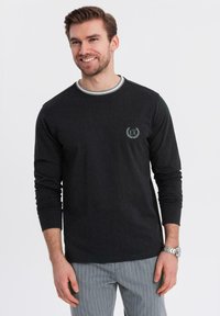 Schwarzes Langarm-T-Shirt mit einem runden, grau besäumten Ausschnitt, das ein kleines Logo auf der Brust trägt. Kombiniert mit gestreiften Hosen.