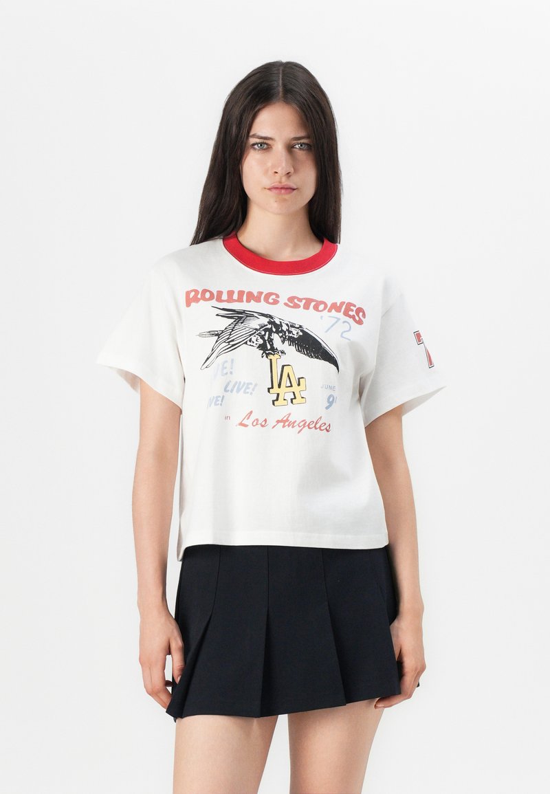 White cotton t-shirt with a red ribbed collar, featuring a graphic print of an eagle, text, and "LA" in yellow. Short sleeves, relaxed fit.