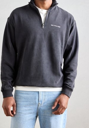 Mężczyzna w ciemnoszarej bluzie z półguzikiem z logo "MARC O'POLO DENIM" i jasnoniebieskich dżinsach na jednolitym tle.