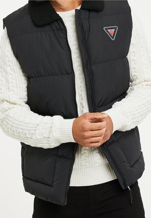 Homme portant une veste matelassée noire avec fermeture éclair et col par-dessus un pull blanc en maille torsadée, les mains jointes devant.