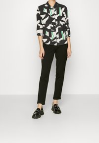 Chemise à motifs noir et blanc avec accents verts, manches trois-quarts, associée à un pantalon noir ajusté et des mocassins noirs à semelles épaisses.