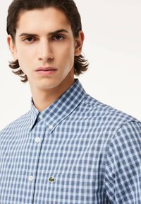 Camisa de cuadros azul y blanca, de botones, con cuello clásico, mangas cortas y un pequeño logo verde bordado en el bolsillo del pecho.
