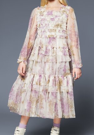 Fille portant une robe florale à manches longues et à volants dans des tons doux rose et beige, assortie à des chaussettes violettes et des baskets blanches.