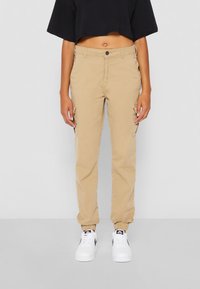 Beige cargobyxor med elastiska muddar, två sidofickor och knäppning, tillsammans med vita sneakers. Toppens är svart och kort. 