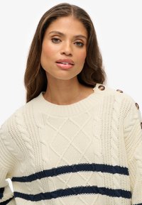 Cremfarbener Kabelstrickpullover mit navyblauen horizontalen Streifen. Mit rundem Halsausschnitt und Knopfakzenten an den Schultern. Strukturiertes Muster über das ganze Kleidungsstück.