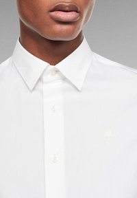 Valkoinen nappikauluspaita, jossa on levitetty kaulus ja hienostunut rakenne. Paidassa on kirjailtu logo toisen napin yläpuolella.