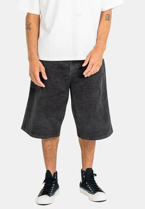 Man wearing knee-length black corduroy shorts, white t-shirt, and black high-top sneakers standing against a plain background.