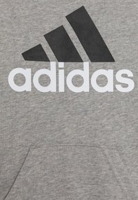 Tissu gris avec le logo à trois bandes noirs d'Adidas et le texte blanc 'Adidas' imprimé au-dessus d'une poche frontale.