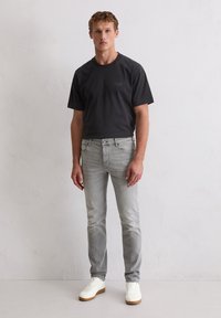 Jeune homme debout contre un mur blanc portant un t-shirt noir à manches courtes, un jean slim gris clair et des baskets blanches.