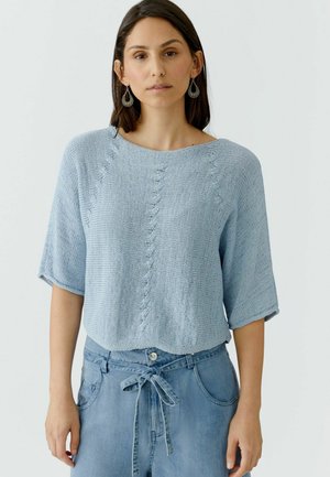 Frau trägt einen hellblauen, kurzärmeligen Strickpullover und blaue Jeans mit gebundener Taille, steht vor einem schlichten, hellen Hintergrund.