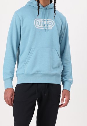 Osoba w jasnoniebieskiej bluzie z kapturem marki Nike z białym logo oraz czarnych spodniach dresowych Nike, stojąca na tle jednolitego białego tła.