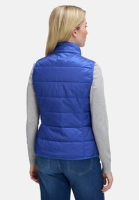 Blaue, gepolsterte Weste mit hohem Kragen und horizontaler Steppung. Über einem grauen Langarmshirt getragen. Glatte Textur, leichtes Material.