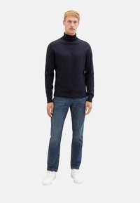 Maglione a collo alto navy in morbido materiale a maglia, abbinato a jeans blu e sneakers bianche; design aderente, maniche lunghe, senza motivi visibili.
