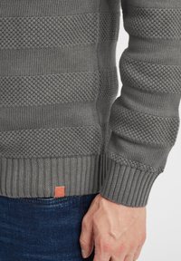 Suéter gris de punto con patrones de franjas texturizadas, puños y dobladillo acanalados, y una pequeña etiqueta de cuero con la marca en la parte inferior.