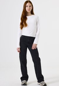 Langärmliges, weißes Oberteil mit kleinen Verzierungen, kombiniert mit dunklen, gemusterten Hosen. Modell trägt hellfarbige Sneakers.