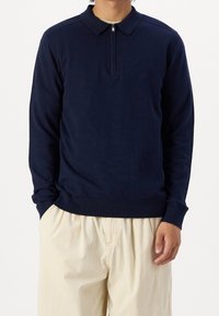 Polo en laine bleu marine à manches longues avec col zippé, poignets et ourlet côtelés, en tissu lisse, accordé avec un pantalon de couleur claire.