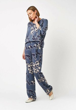 Woman standing, wearing a blue floral patterned loose top and wide-leg pants with white flat shoes against a plain white background.