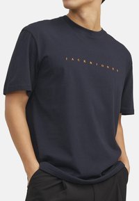 T-paita tummansinisestä puuvillasta, lyhyillä hihoilla, jossa on "JACK & JONES" tulostettuna oranssilla etupuolella, pyöreä pääntie ja rento malli.
