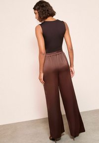 Schwarzes ärmelloses Oberteil kombiniert mit hochtaillierten, weit geschnittenen, braunen Satin-Hosen, die über einen elastischen Bund und eine glatte Textur verfügen.