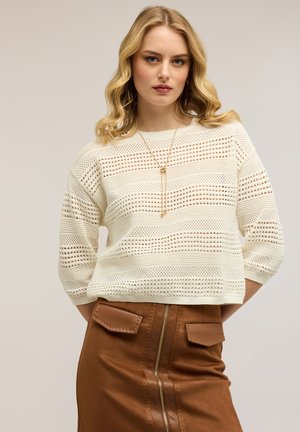 Frau mit welligem blondem Haar, die einen weißen gestrickten Pullover und einen braunen, hoch taillierten Rock mit Reißverschluss vorne und Taschenklappen trägt.