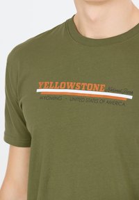 Oliven grøn bomulds t-shirt med teksten "YELLOWSTONE National Park" i orange, hvid og sort. Rund halsudskæring med korte ærmer.