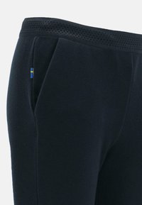 Fleece-housut tummansinisestä kankaasta, jossa on teksturoitu vyötäröosa ja verkkomateriaalia. Sisältää pienen sinisen ja keltaisen lippuaccentin.