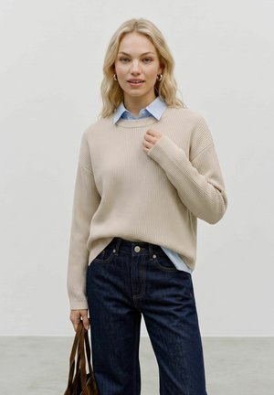 Lyshåret kvinde iført en beige sweater over en blå skjorte med krave, mørke jeans, holder en brun lædertaske, står foran en ensfarvet baggrund.