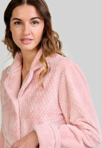 Mujer con cabello ondulado castaño claro viste un albornoz suave de color rosa texturizado frente a un fondo gris claro liso.