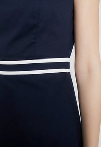 Marinblå klänning med en åtsittande överdel och en vit dubbelrand runt midjan. Slät tyg med en lätt glans, strukturerad design.