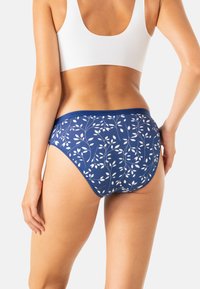 Femme portant un sous-vêtement bleu avec un motif de feuilles blanches et un soutien-gorge de sport blanc, se tenant devant un fond clair.