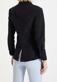 Donna che indossa un blazer nero aderente a maniche lunghe con uno spacco sul retro, abbinato a pantaloni aderenti di colore azzurro, in piedi davanti a uno sfondo neutro.