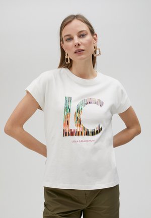 Camiseta blanca de algodón con un gráfico colorido de "LC" hecho de diseños a lápiz, con mangas cortas y ajuste relajado. Combinada con pantalones verde oliva.