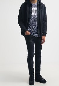 Černá kapucní bunda přes vzorovanou košili, spárovaná s tmavými skinny džíny a černými teniskami. Zaměření na vrstvy a kontrastní textury.