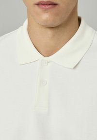 Polo shirt blanc avec un col plat, une patte de boutonnage à trois boutons et un tissu doux et texturé. Design simple sans motifs ni accents.