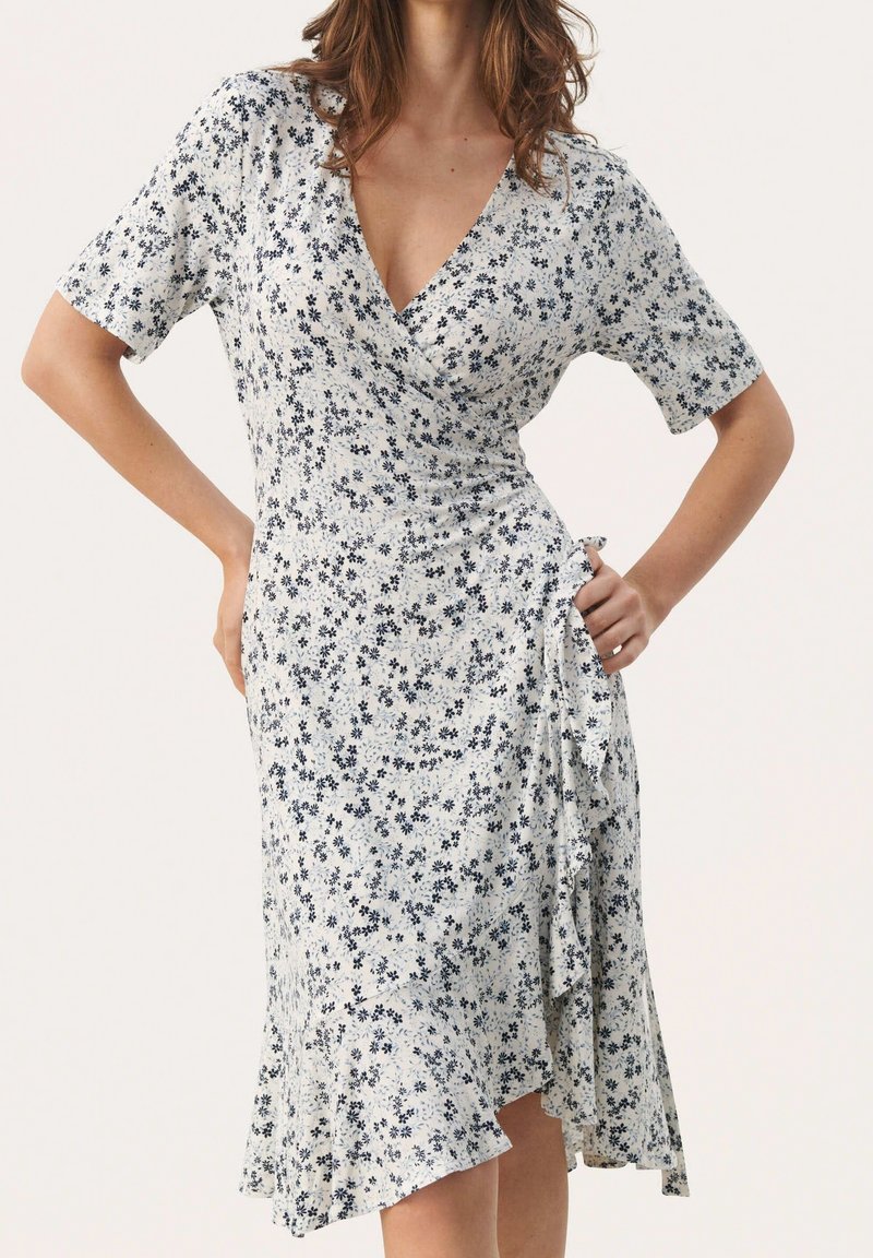 Femme portant une robe portefeuille blanche à manches courtes avec un petit imprimé floral noir, posant les mains sur les hanches devant un fond uni.