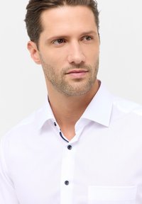Camicia bianca con colletto, realizzata in tessuto liscio, con bottoni blu scuro e una tasca sul lato sinistro.
