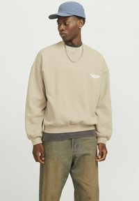Beige sweatshirt with crew neckline, loose fit, and ribbed cuffs. Accented with a small white logo on the chest. Paired with blue cap and denim pants.