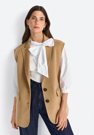 Steppweste aus Kamelhaar mit taillierter Passform, Reverskragen, zwei Taschen vorne und zwei schwarzen Knöpfen. Kombiniert mit einer weißen Bluse und dunklen Jeans.