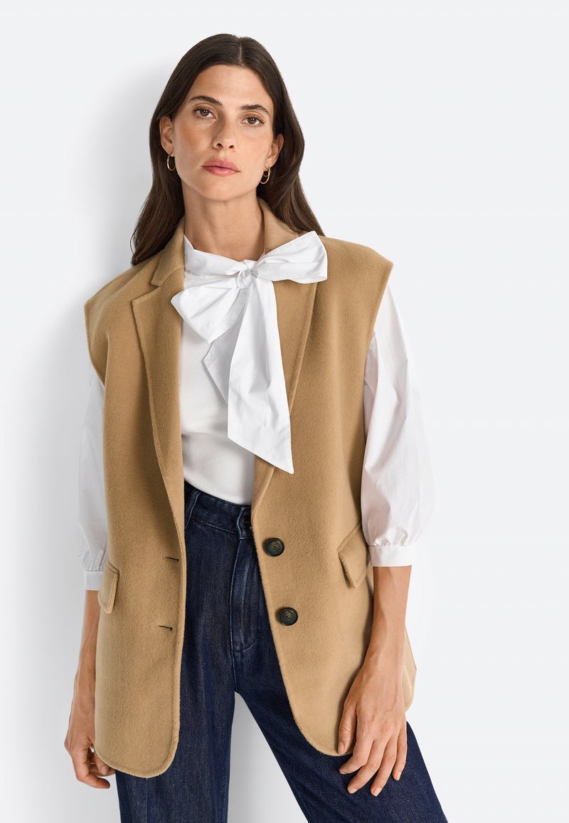 Camel kleurige wollen vest met een gepersonaliseerd ontwerp, met notch revers, twee voorknoopzakken en twee zwarte knopen. Gepaard met een witte blouse en donkere spijkerbroek.