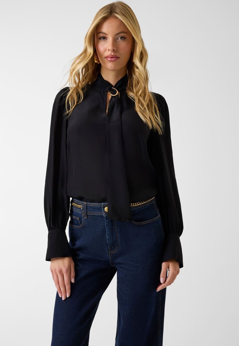 Femme blonde portant un chemisier noir à manches longues avec un col noué et un détail de bague dorée, assorti à un jean bleu foncé et une ceinture chaîne.