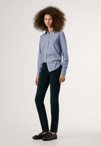 Modra in bela prugasta srajca s klasičnim ovratnikom, v paru s temnimi jeans hlačami in rjavimi loaferji, ki imajo okrasne žebljičke.