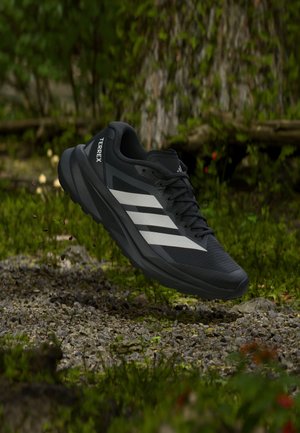 Černý běžecký terénní bot Adidas Terrex s bílými pruhy vznášející se nad štěrkovou cestou v lesním prostředí.