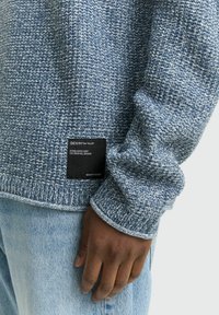 Blauwe gebreide trui met een gestructureerd patroon en geribbelde manchetten. Bevat een zwart merklabel aan de onderzijde. Draagt met lichtblauwe jeans.