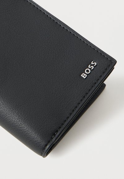 Portefeuille bifold en cuir noir avec des coutures fines et le logo argenté de la marque BOSS dans un coin, posé sur une surface claire et unie.