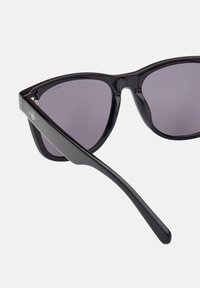 Gafas de sol de plástico negro con diseño de ojo de gato, lentes oscuros, acabado brillante y detalle del logo en la patilla.