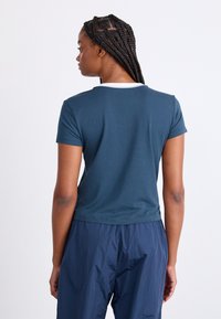 T-shirt κοντομάνικο, στενής γραμμής σε σκούρο τιρκουάζ, με λευκή επένδυση στο λαιμό. Το ύφασμα φαίνεται λείο με ελαφριά ελαστικότητα, φορεμένο με ναυτικές παν trousers.