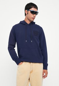 Navy blue sweatshirt with a front pocket, drawstring hood, and tonal logo. Paired with light-colored jeans and sunglasses.